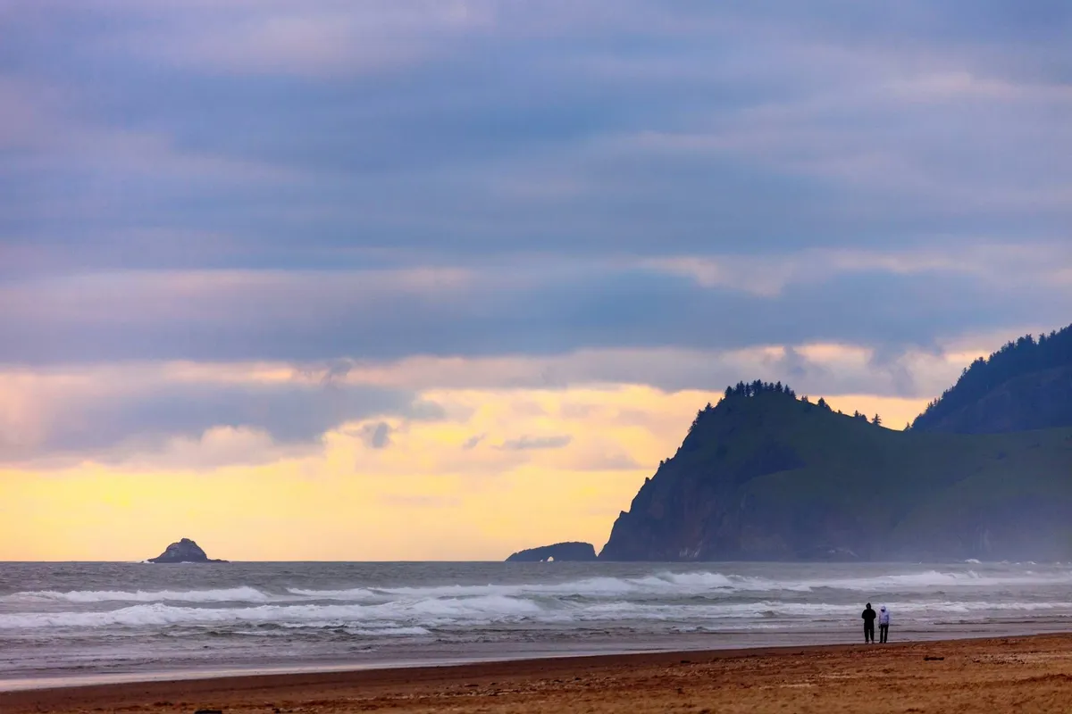 A wide, picturesque stretch of beach at sunset, with gentle waves, distant cliffs, and a couple walking along the shore under a glowing sky—evoking a sense of tranquility and adventure right at the doorstep.