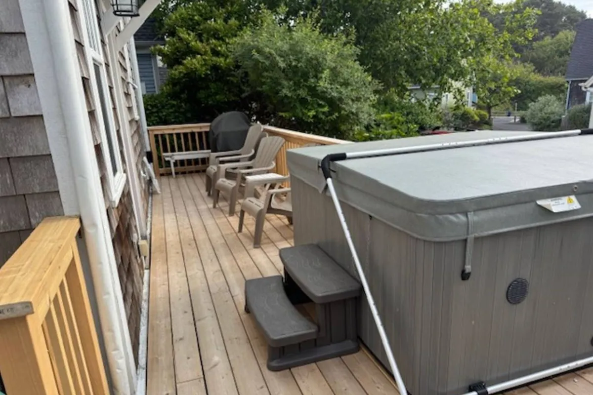 A view of the wooden deck featuring a bubbling hot tub, comfortable outdoor seating, and glimpses of lush greenery in the background. The scene is bathed in soft evening light, suggesting the approach of sunset and an atmosphere perfect for relaxation or stargazing.