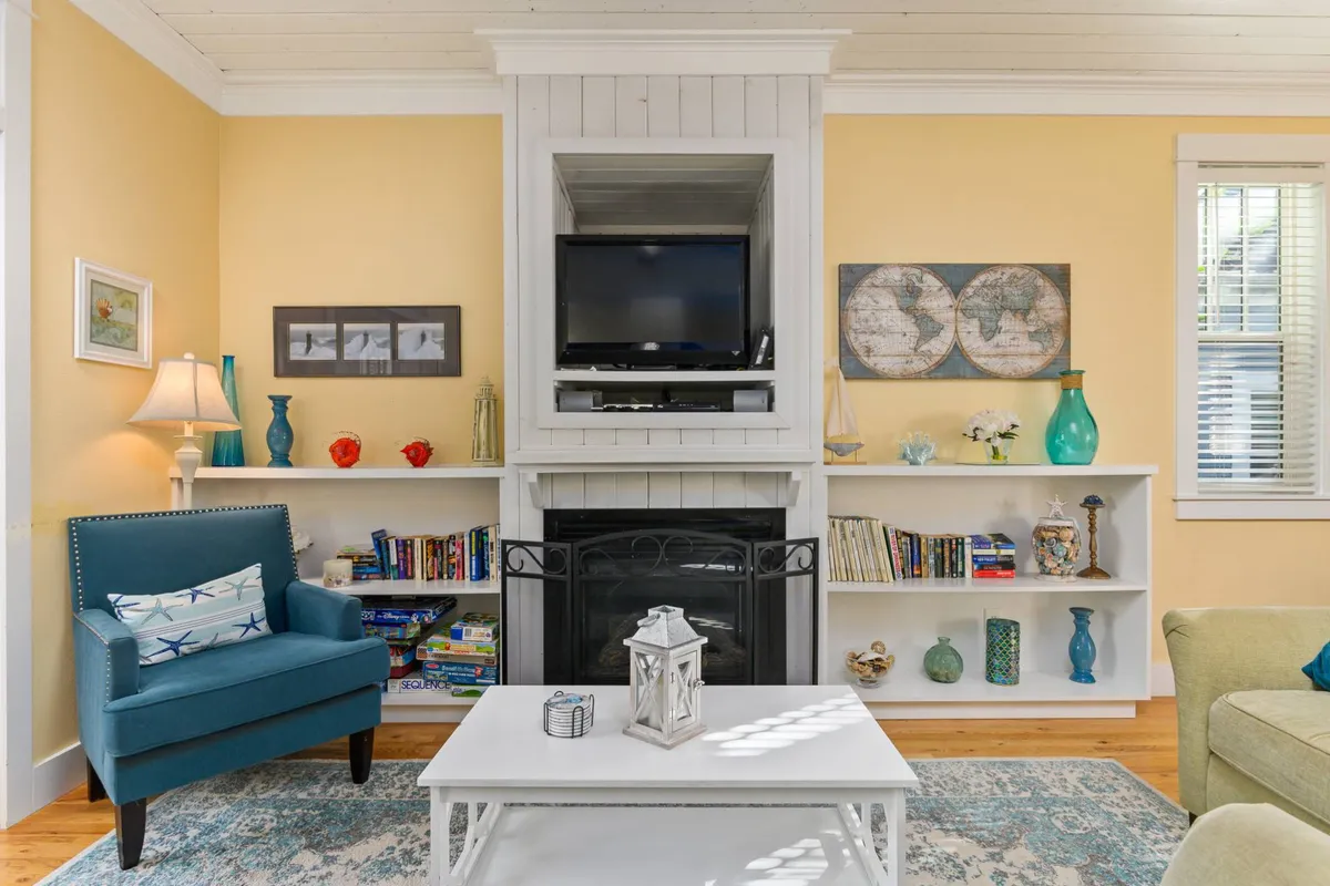 A bright and cozy sitting area highlighted by a blue armchair near a central natural gas fireplace. The space includes a coffee table and a television, with ample natural light filtering in, making it ideal for both relaxing mornings and lively gatherings.