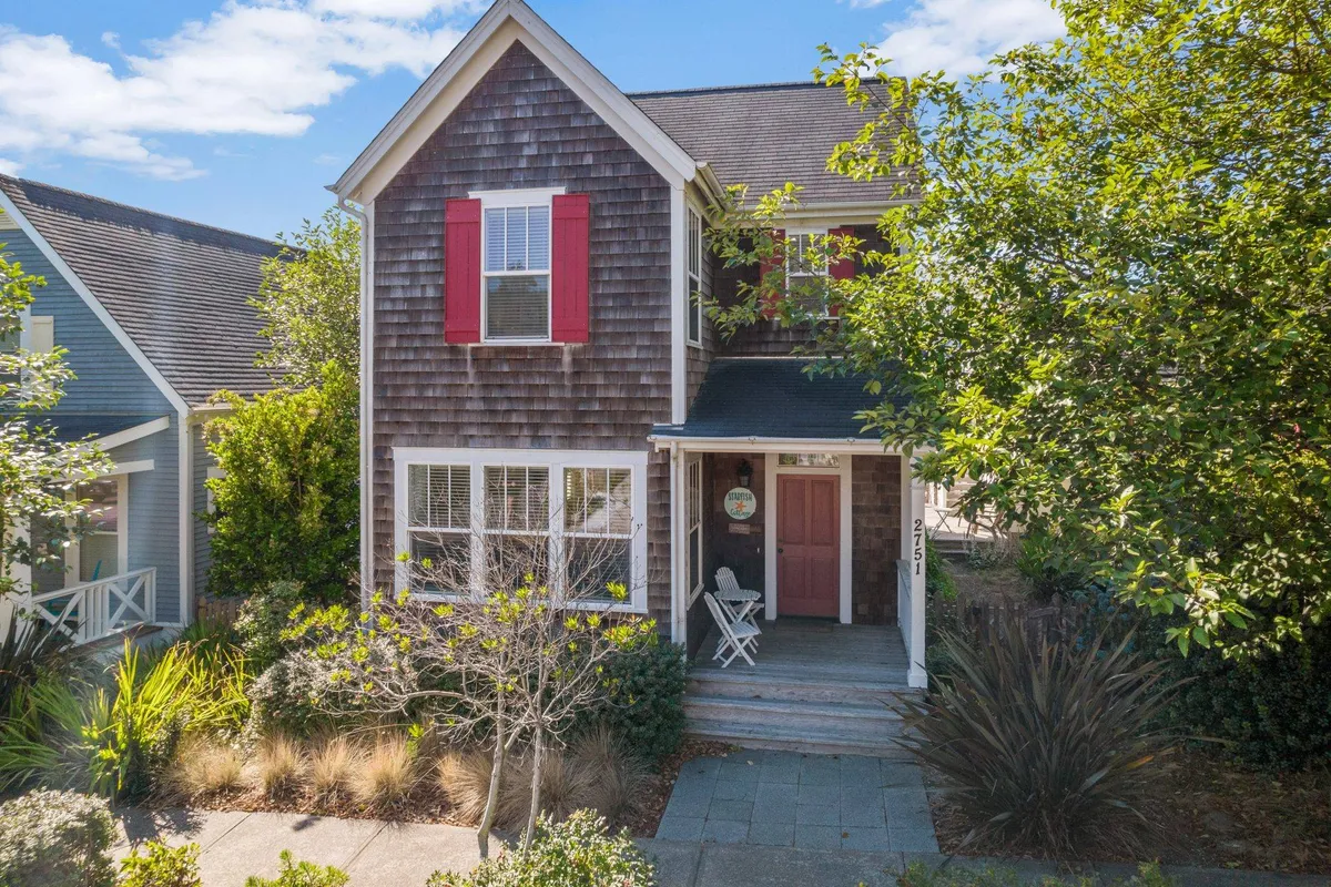 A welcoming exterior view of Starfish Cottage, showcasing its charming wooden shingle facade, vivid red shutters, and a front porch with inviting seating surrounded by lush, blooming greenery.