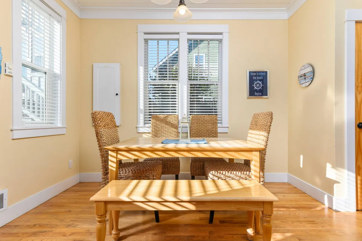 A close-up of the dining table, surrounded by woven chairs and accented with nautical wall décor. Natural light pours in from a nearby window, creating a cozy, relaxed ambiance for meals or conversation.