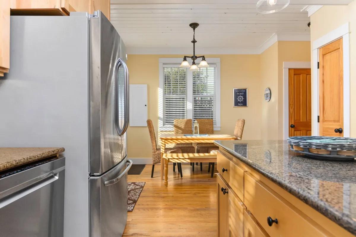 A bright, open dining area with a granite countertop visible in the kitchen, a stainless steel refrigerator, and a dining table set with wicker chairs. The setup encourages communal meals, with a welcoming and family-friendly atmosphere.
