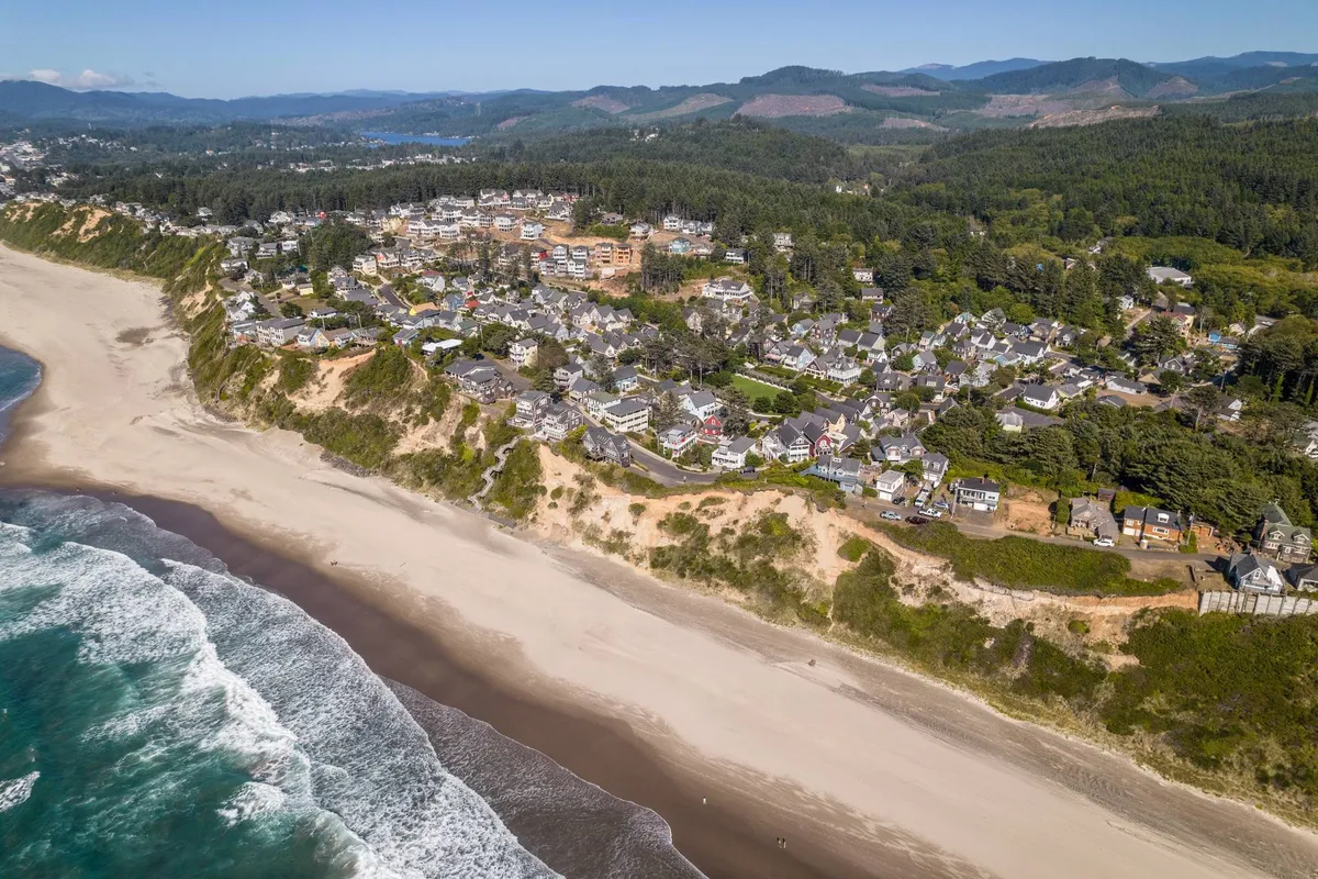 A sweeping view of the Oregon Coast with sandy beach, rolling ocean waves, dramatic cliffs, and a line of forest in the background. The natural beauty of the coastline is prominent with space for outdoor activities and relaxation.