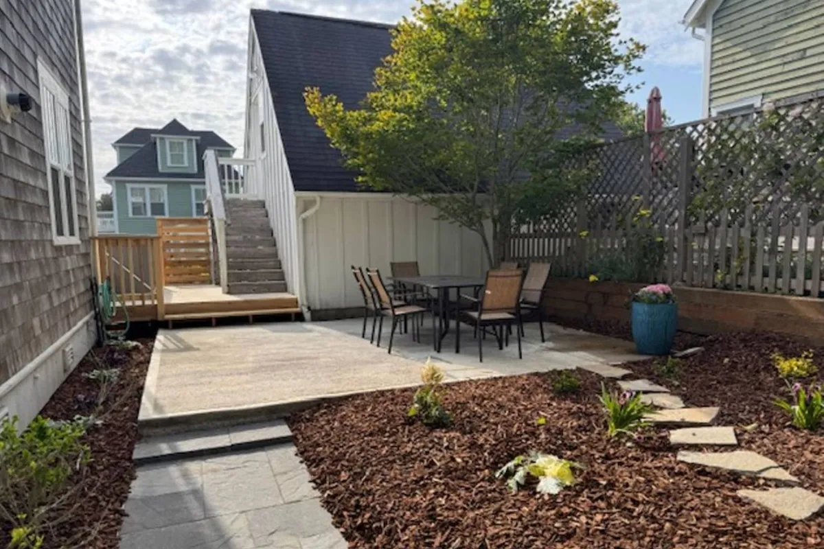 An outdoor patio dining area set beneath a leafy tree, featuring a dining table and chairs surrounded by tranquil garden landscaping and decorative planters, creating an ideal setting for al fresco meals or casual gatherings.