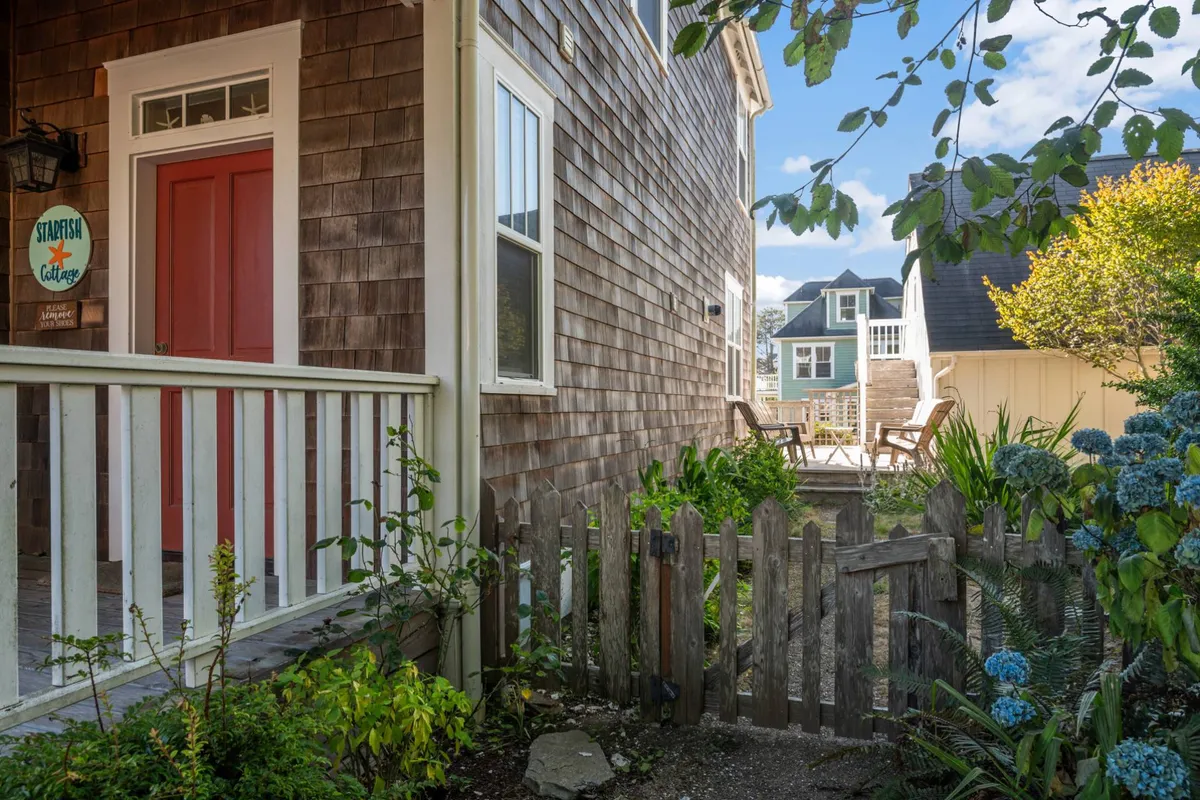 This exterior image highlights the front of Starfish Cottage, featuring a welcoming red door, a wooden fence, lush greenery, and blooming blue flowers. The inviting front porch showcases comfortable seating, creating a romantic and serene exterior ambiance.