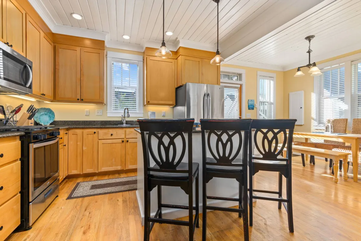 A warmly lit kitchen centered around an island with bar stools, featuring stainless steel appliances and wooden cabinets. The layout is open and inviting, ideal for cooking and social interaction, with ample space for meal preparation and gathering.