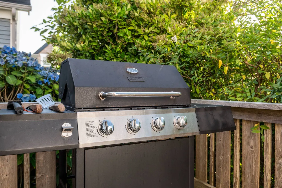 An outdoor grill is set up in a green space, surrounded by trees and plants, with all the essentials for barbecuing. The setup hints at alfresco dining and festive gatherings, with a relaxed, inviting vibe.
