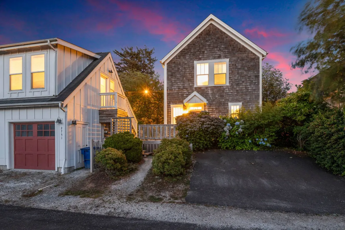 An exterior view showing two houses with distinct architecture, illuminated windows, and lush landscaping. The setting evokes a neighborhood ambiance, suggesting proximity to nature walks and local attractions.