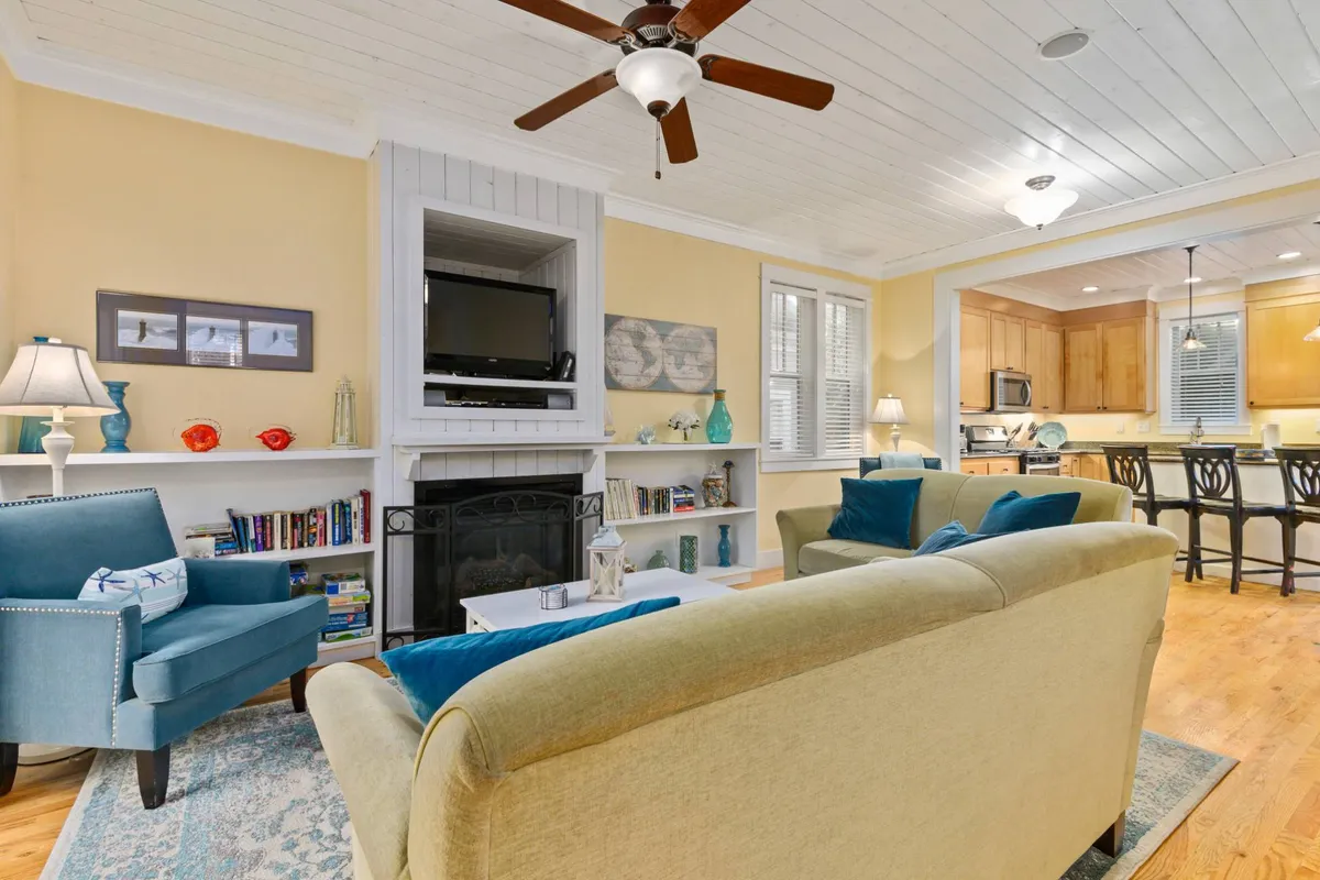 A cozy living area featuring a plush sofa, a blue armchair, a glowing gas fireplace, and a bookshelf. The adjacent kitchen area is visible in the background, creating an open, sociable layout. Warm lighting and soft textiles enhance the welcoming, comfortable mood.